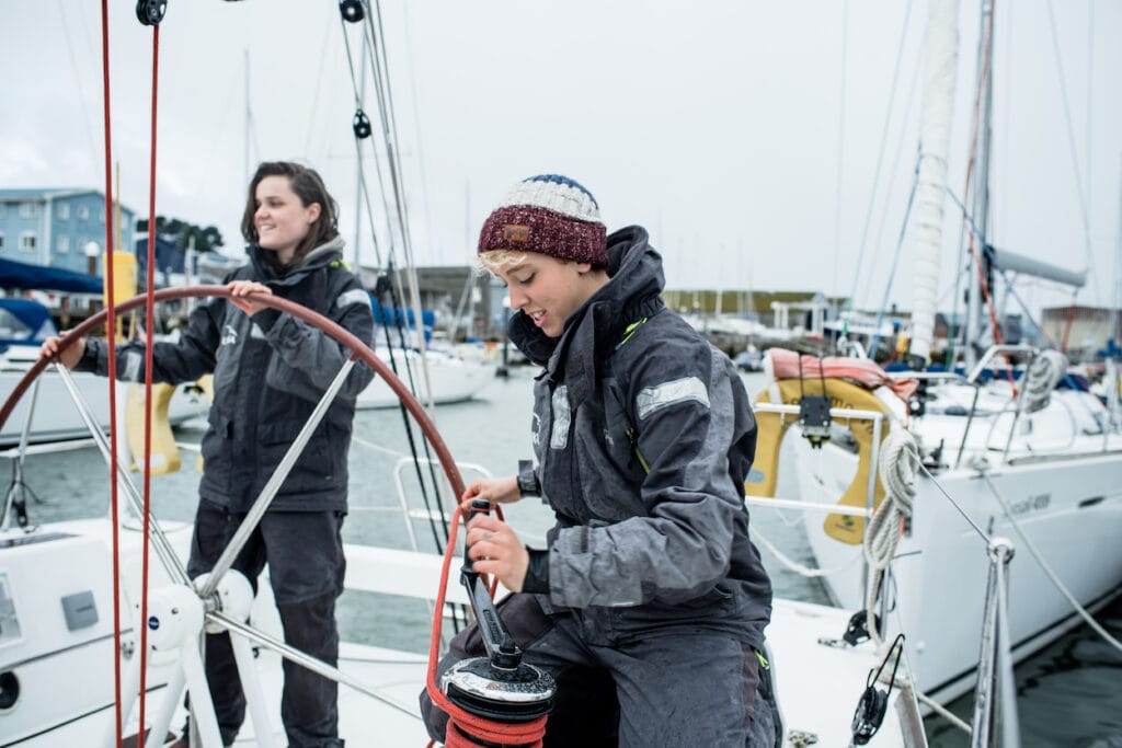 A person at the helm of a yacht with another working a winch nearby, whilst starting a manoeuvre out of a marina mooring.