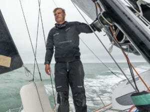 Man standing on deck of racing sailing boat dressed in high-performance gear.