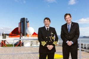 Two men, one in merchant navel uniform and the other in a dark suit, stand on the top deck of a cruise liner