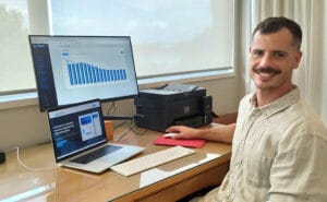 man with a moustache sitting at a desk with a laptop and second screen