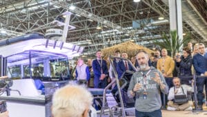 white male speaking to crowd with boat behind in exhibition hall