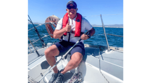 A sailor gives a jolly thumbs up from a boat's stern, with the sea stretching into the distance