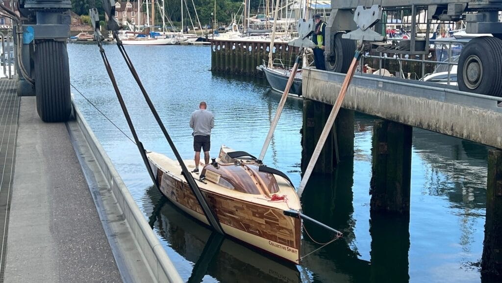 Boat made of 1200 wooden items lowered into marina berth while man stands on deck. Collective Spirit was Olympic art project.