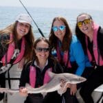 Four women in fishing boat with pink lifejackets on one holding a caught fish