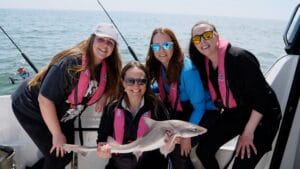 Four women in fishing boat with pink lifejackets on one holding a caught fish