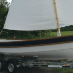 The hull of a boat on a trailer with sails up. It's a Drascombe Gig as ordered by the French Navy