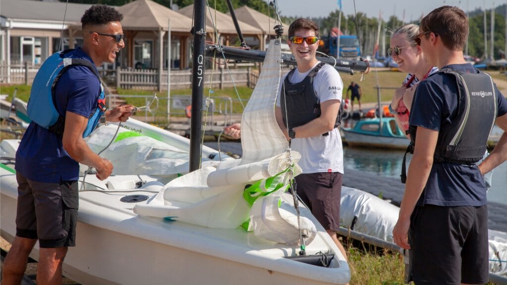 Four sailors stand around dinghy laughing