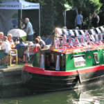 canal boat decorates with UK flags docked alongside a party of people at a table. This is Ware Boat Festival cancelled due to towpath being dangerous