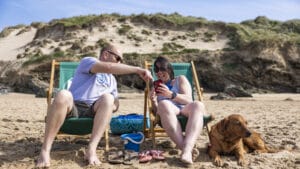 Two people sit in deckchairs on the beach using a mobile phone.