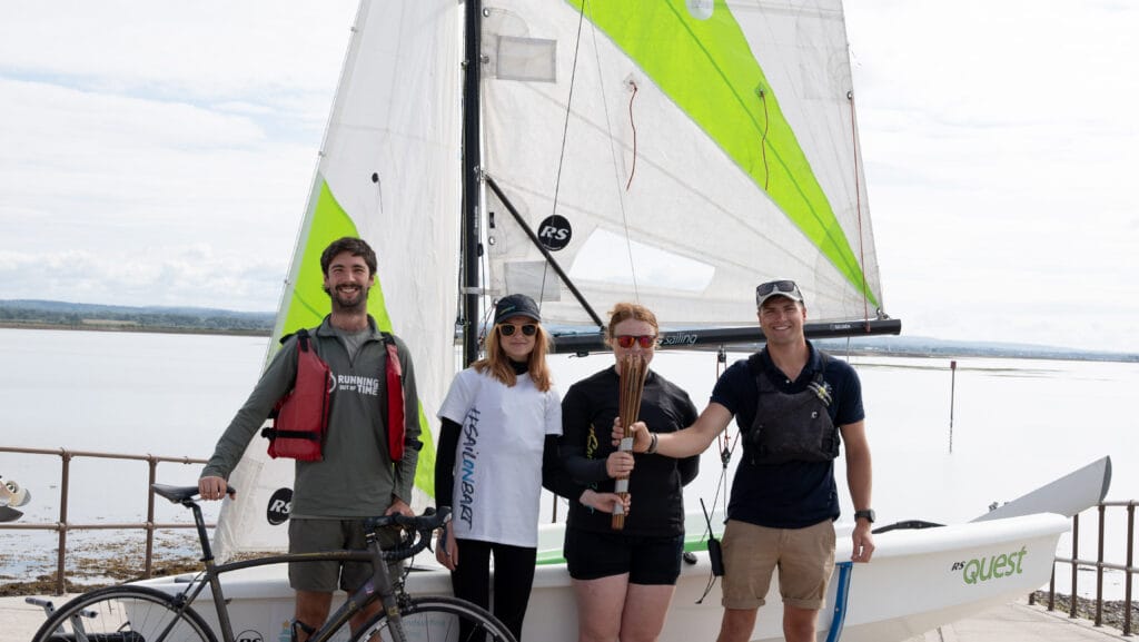 People gather in front of a small sailboat with white and green sail.