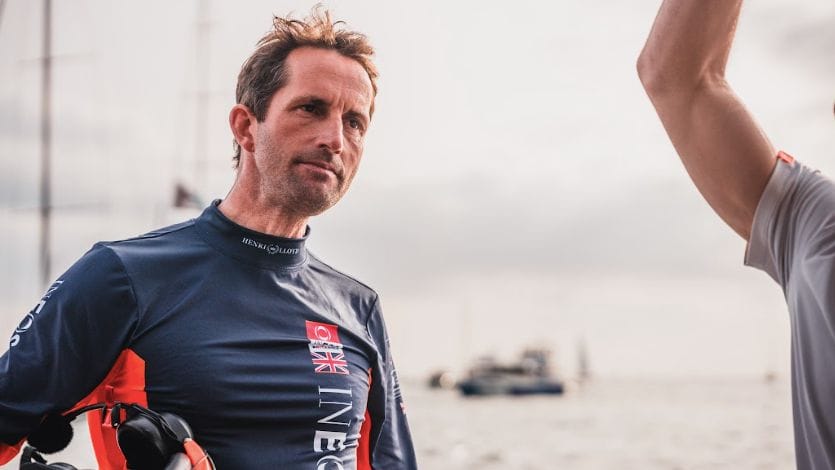 Ben Ainslie looks away from camera. He stands with boats in background.