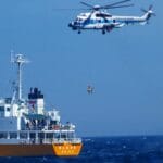 Helicopter hovers as it rescues a woman from the ocean, with a ship nearby.