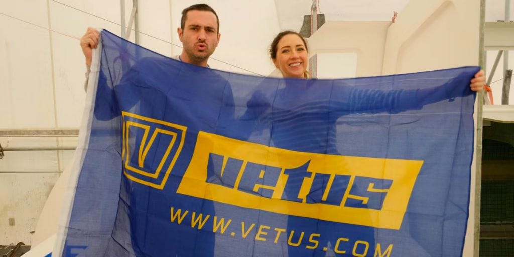 A couple smile at the camera from behind a blue and yellow Vetus flag. Vetus is sponsoring MJ Sailing as part of its marine marketing mix
