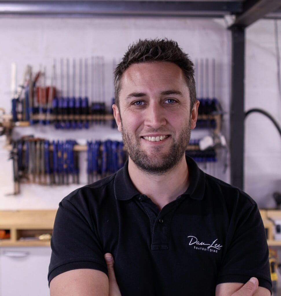 Dan Lee smiles at camera in front of a stack of boat building tools