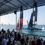 A crowd goes wild on shore as it watches SailGP boats compete. Image from back, over crowd's head.