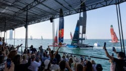 A crowd goes wild on shore as it watches SailGP boats compete. Image from back, over crowd's head.