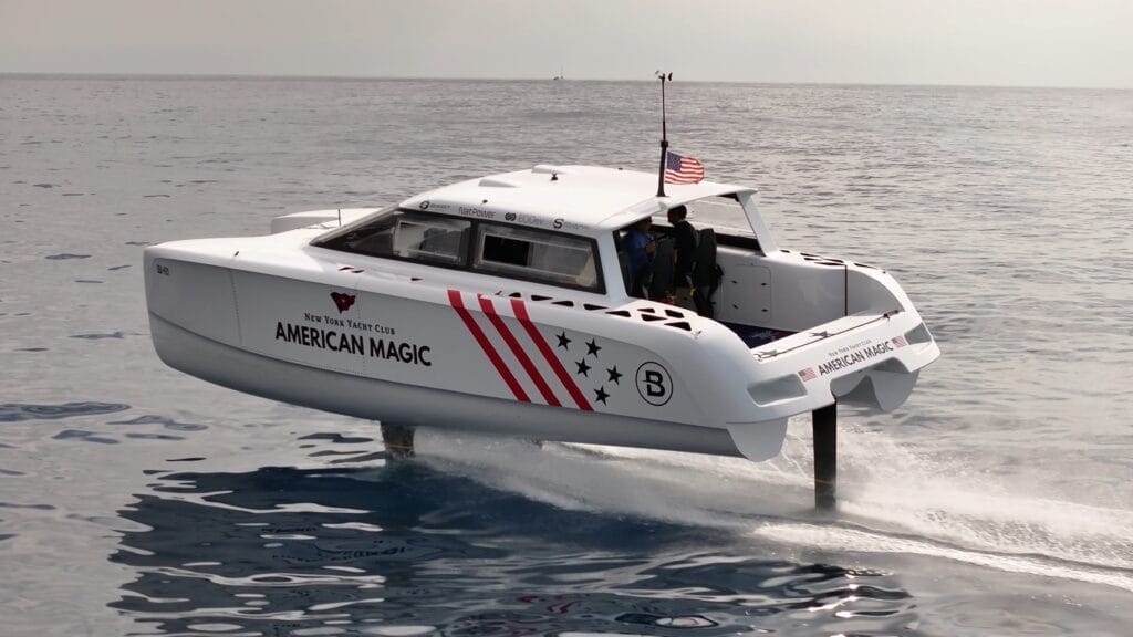 Motorboat on foils sweeps across sea with American Magic branding. This is Bluegame boat.