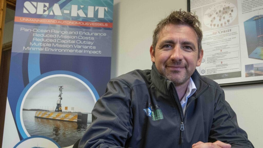 Man smiles at camera in from of a picture of an uncrewed vessel on the water